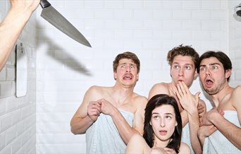 Three men, one woman in towels in a shower someone is holding a knife up out of shot and they look shocked.