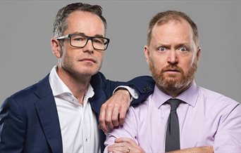 Two white men, one wearing glasses with a shirt and jacket. The other has a beard with a shirt and tie.