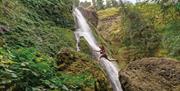 Green landscape with a waterfall in the middle and a man jumping from a rock to a ledge.