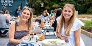 Two ladies enjoying food and drinks on outdoor benches at the Queens Hotel