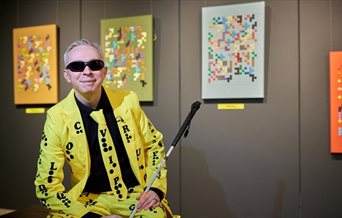 Journey By Dots, Clarke Reynolds - featuring the artist in a bright suit sat in front of some of his pieces