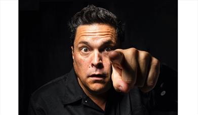 Press shot of Dom Joly in black pointing at the camera