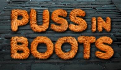 Logo for Puss in Boots at Groundlings Theatre, featuring the show name in ginger furry letters, against a background of a cobbled street