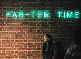 Lady stood in front of brick wall with neon sign: par-tee time