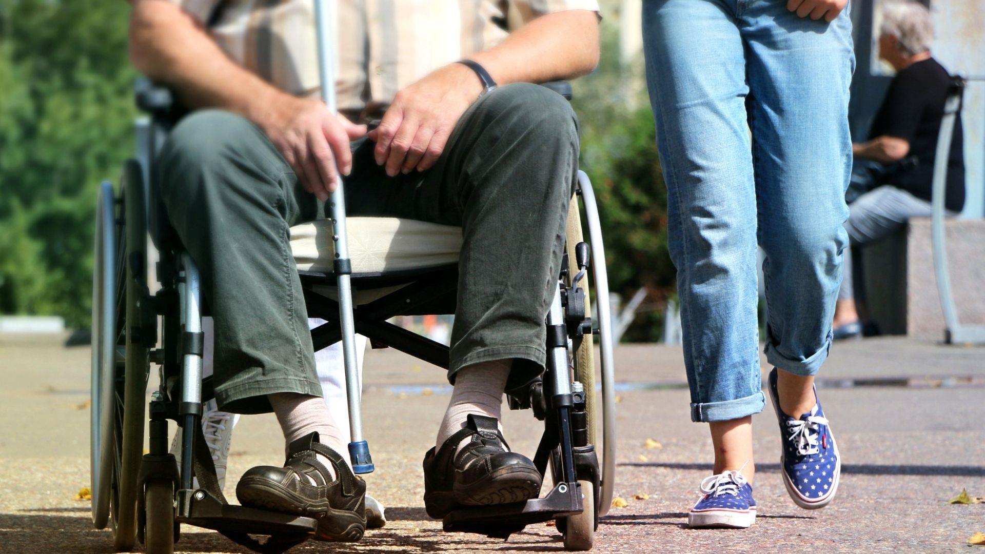 Man in wheelchair holding walking stick, with lady walking by his side.