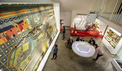 View from above of visitors standing around a table play an interactive game whilst look at a huge glass case containing a large fabric wall-hanging