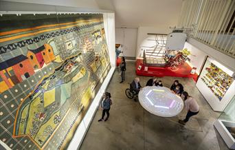 View from above of visitors standing around a table play an interactive game whilst look at a huge glass case containing a large fabric wall-hanging