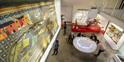 View from above of visitors standing around a table play an interactive game whilst look at a huge glass case containing a large fabric wall-hanging