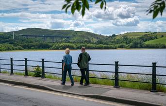 A couple walking along the bank of Hollingworth Lake on a bright day.