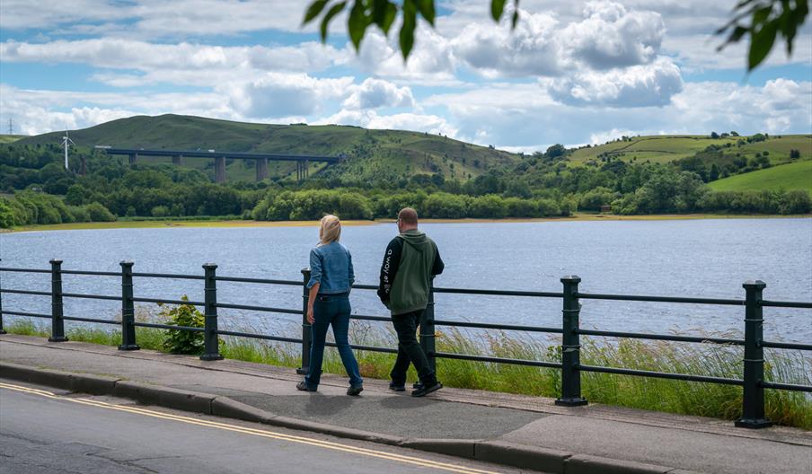 A couple walking along the bank of Hollingworth Lake on a bright day.
