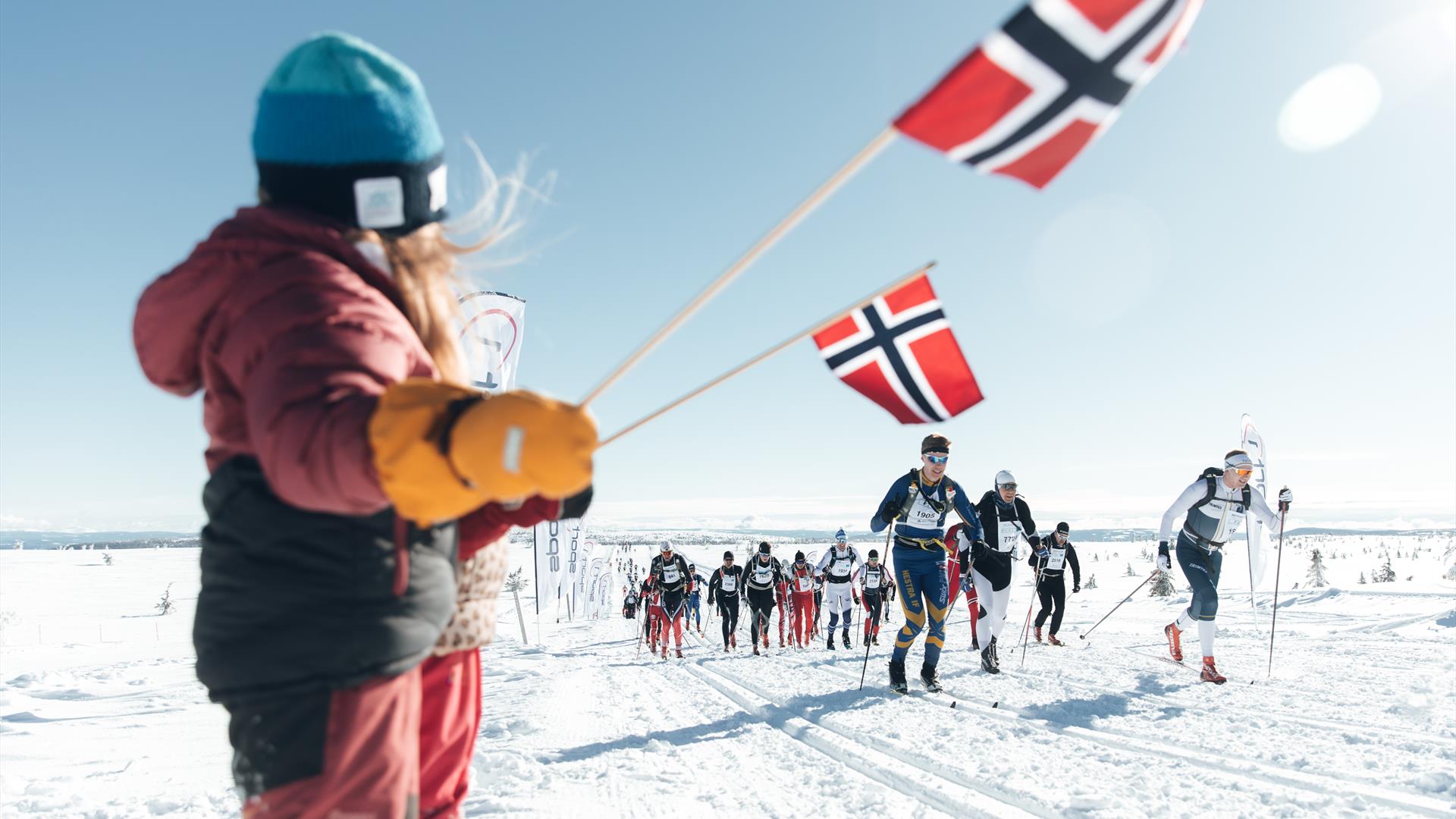 Mange løpere i Birkebeinerrennet kommer i full fart i løypa. På sidelinja viftes det med norske flagg.