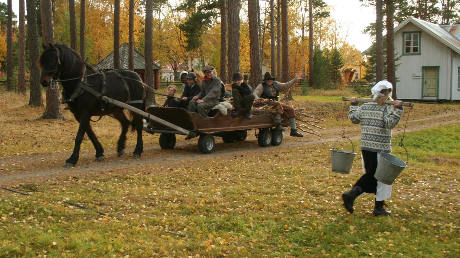 Hest som drar en vogn med en gruppe mennesker. En kvinne bærer to melkespann. Utenfor museumssenteret Ramsmoen.