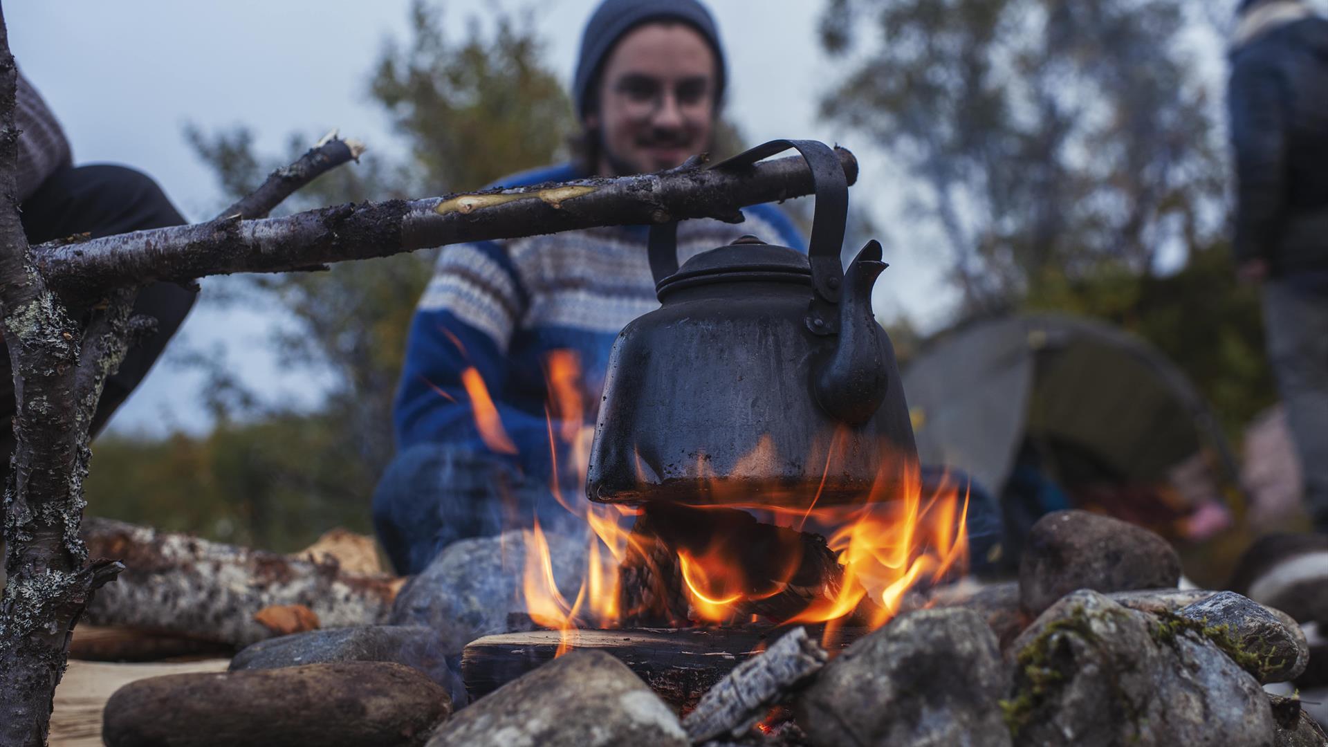 En person sitter ved bålet med svart kaffekjel