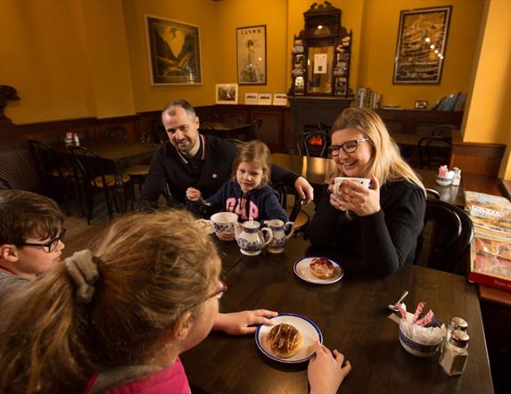 Family of 3 children and 2 adults sitting eating and drinking tea around a wooden dining table in the railway musuem cafe