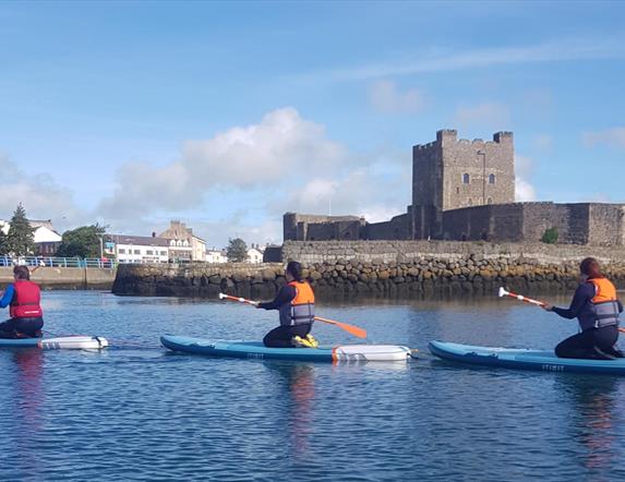 image of 3  paddleboarders kneeling on boards in water in front of Carrickfergus Castle.