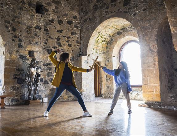 A woman and child having a pretend sword fight in the room at the top of the castle keep with the sun beaming through the window behind.