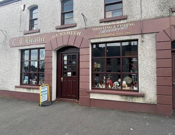 Brown and cream exterior of CF Beattie with sign stating Shooting & Fishing Accessories / Gunsmith plus antique displays in windows