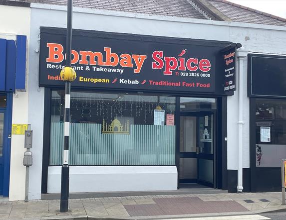 Exterior of Bombay Spice restaurant with pedestrian crossing in front of it and large window with entrance door.