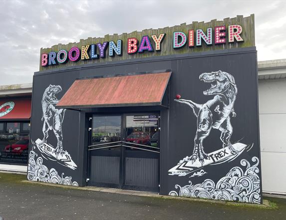 Exterior of Brooklyn Bay Diner entrance with large sign, door canopy and dinosaur wall murals