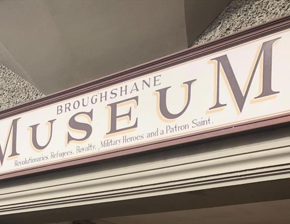 Red and Cream sign on building stating Broughshane Museum - Revolutionaries, Refugees, Royalty, Military Heroes and a Patron Saint.