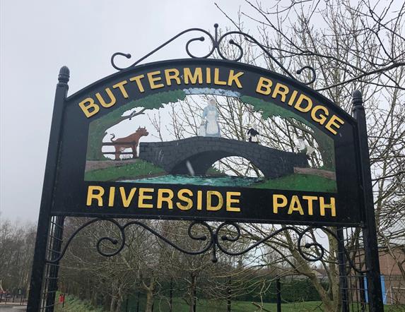 Black metal Buttermilk Bridge Riverside Path sign with writing in yellow and image of a bridge with cow and milk maid crossing