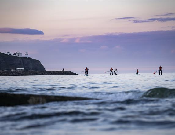 Group of friends trying out paddle boarding in the open water with Blackhead Lighthouse in background