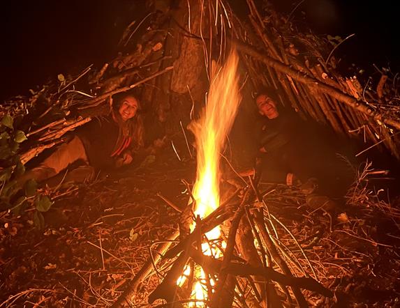 Two women laying under stick huts around a camp fire.