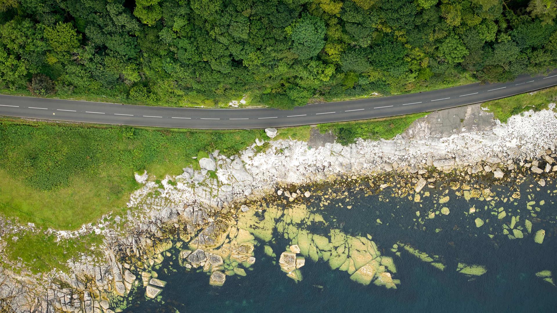 An aerial view of the coast road showing the sea, road and green hills.