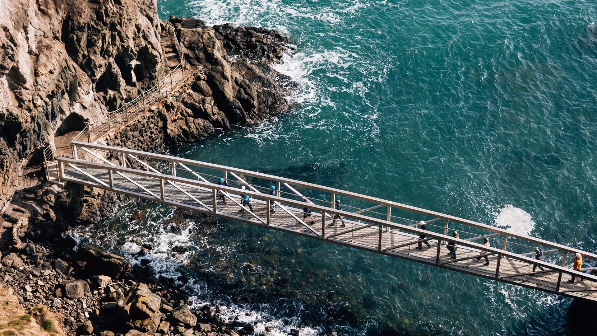An aerial image showing a tour group walking over the first bridge at The Gobbins Cliff Path on a bright day, with sea in background.