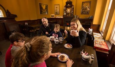 Family of 3 children and 2 adults sitting eating and drinking tea around a wooden dining table in the railway musuem cafe