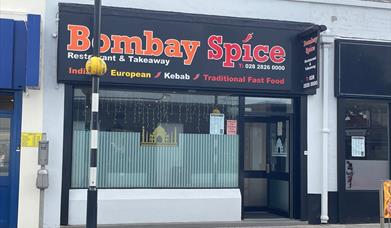 Exterior of Bombay Spice restaurant with pedestrian crossing in front of it and large window with entrance door.