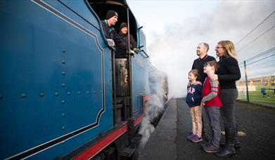 A family of four standing on the platform talking to the driver of a blue steam train 