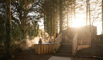A couple perched on the edge of a hot tub elevated above the Forest floor. Views behind them of the River Maine.