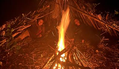 Two women laying under stick huts around a camp fire.