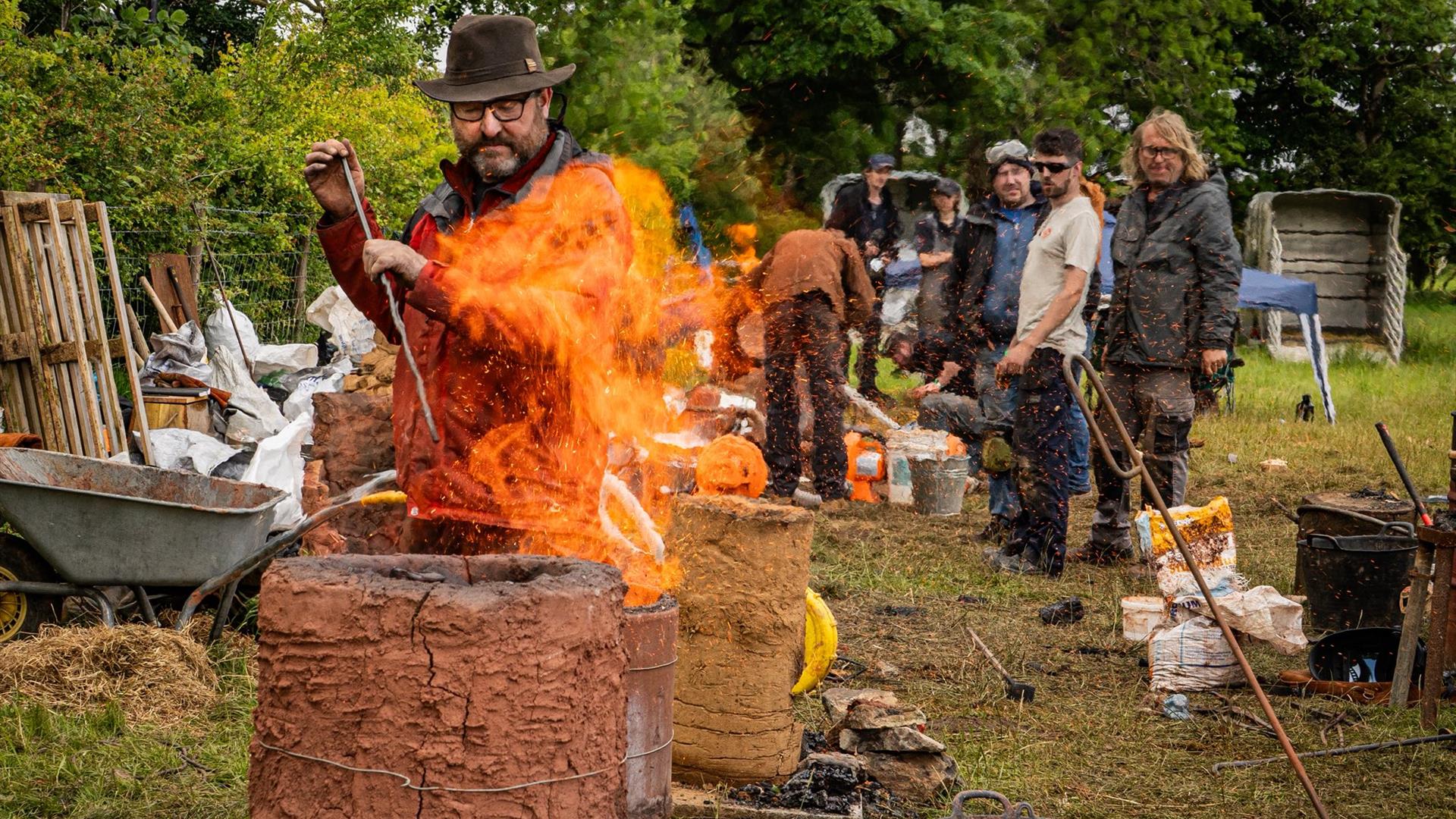 A man tends to a mud furnace with tall flames rising from the top of it, other look on