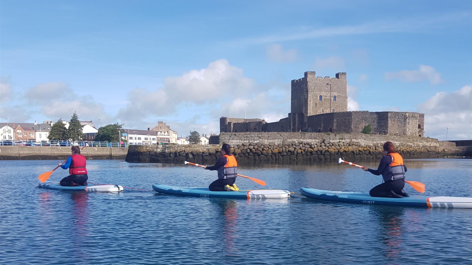 image of 3  paddleboarders kneeling on boards in water in front of Carrickfergus Castle.