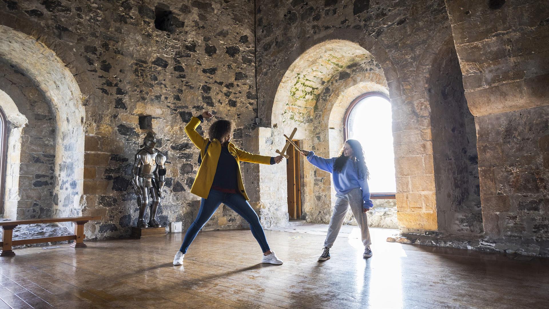 A woman and child having a pretend sword fight in the room at the top of the castle keep with the sun beaming through the window behind.