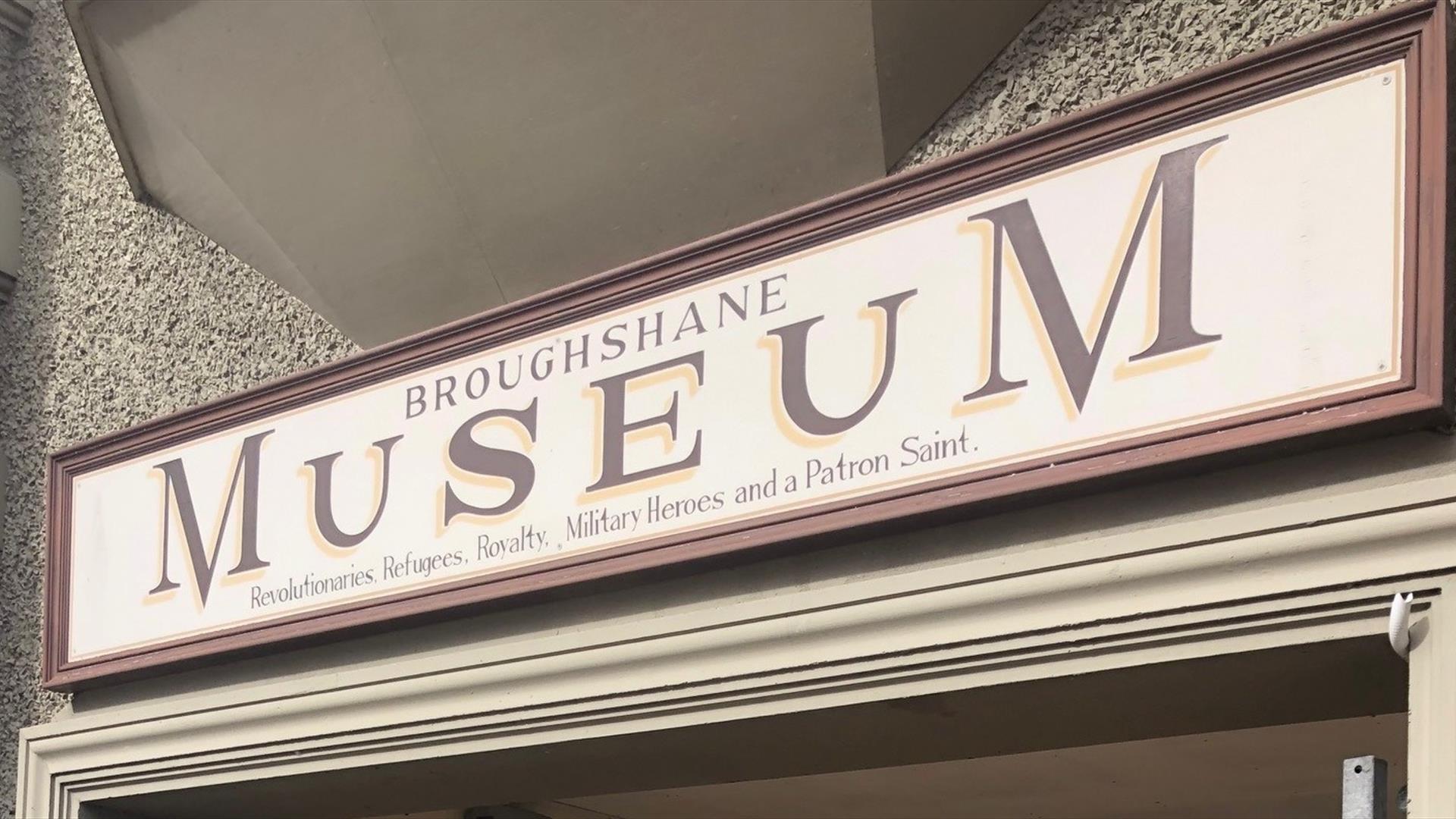 Red and Cream sign on building stating Broughshane Museum - Revolutionaries, Refugees, Royalty, Military Heroes and a Patron Saint.