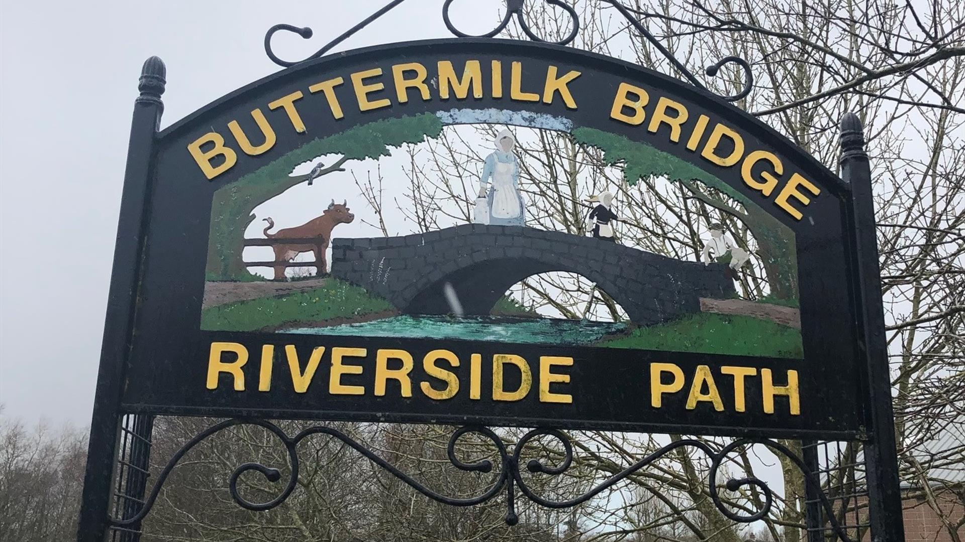 Black metal Buttermilk Bridge Riverside Path sign with writing in yellow and image of a bridge with cow and milk maid crossing