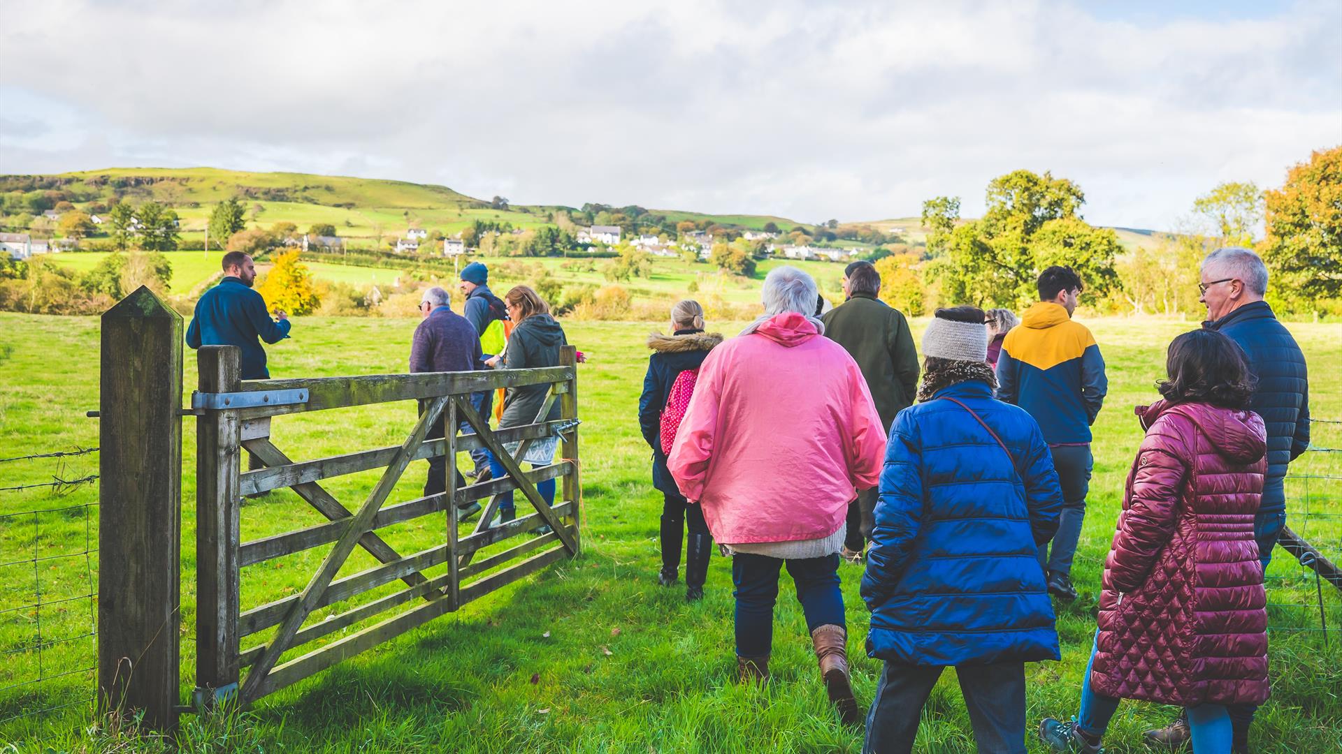 A group of people walking away from the camera, through a wooden gate to a field with a mountain backdrop