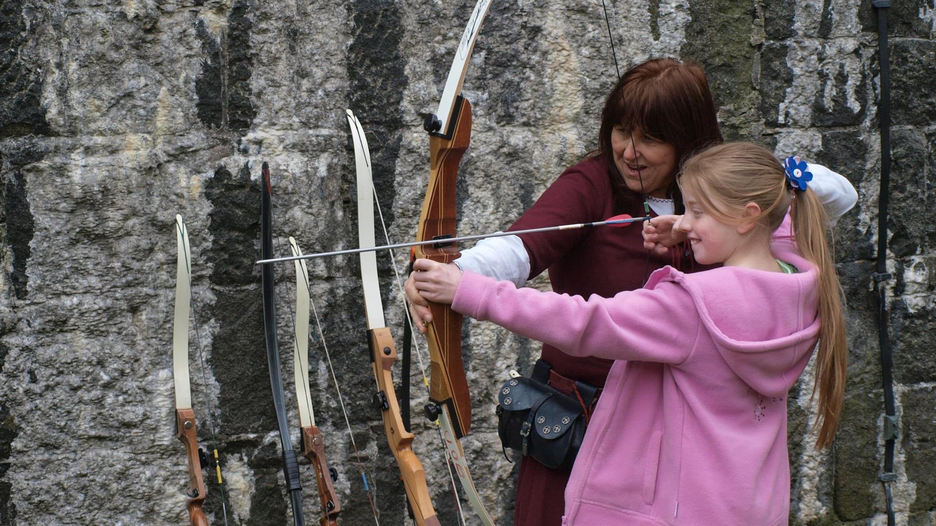 Young girl being shown how to shoot a bow and arrow by female archer in Carrickfergus Castle