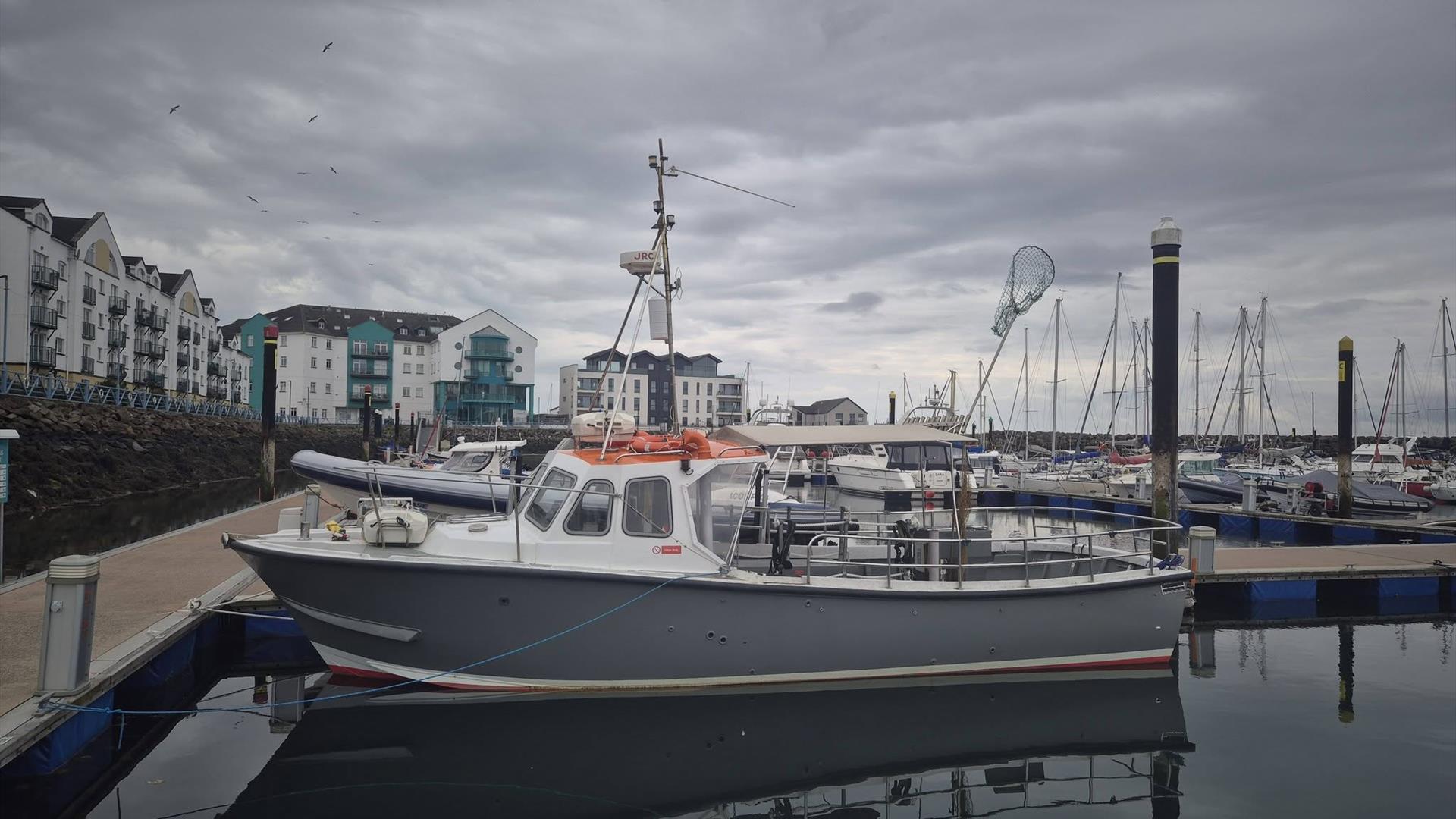 Charter Boats NI's grey and white fishing boat moored at Carrickfergus Marina on a calm day, with modern waterfront apartments, sailing yachts and mar