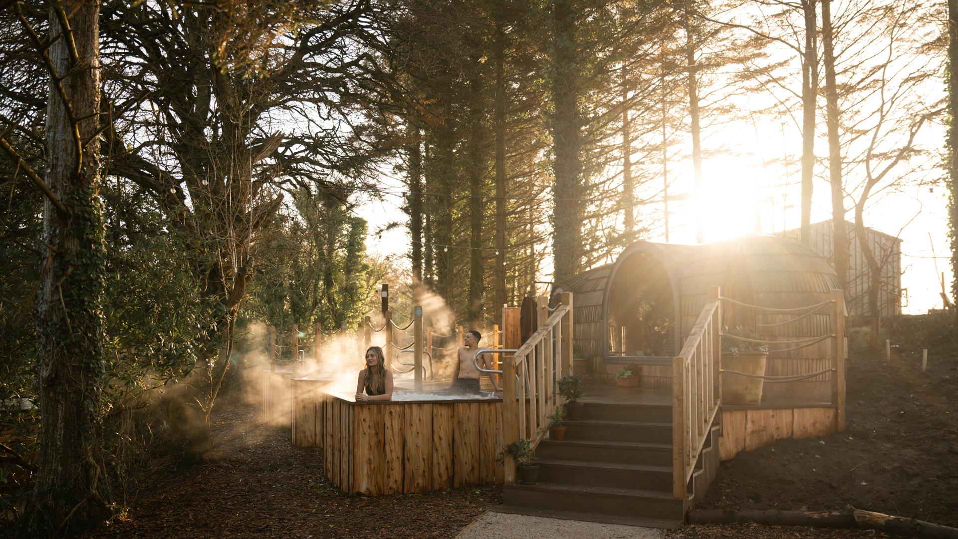 2 guests in outdoor wooden hot tub and barrel‑shaped sauna set among tall trees at sunset, with steam rising from the warm water.