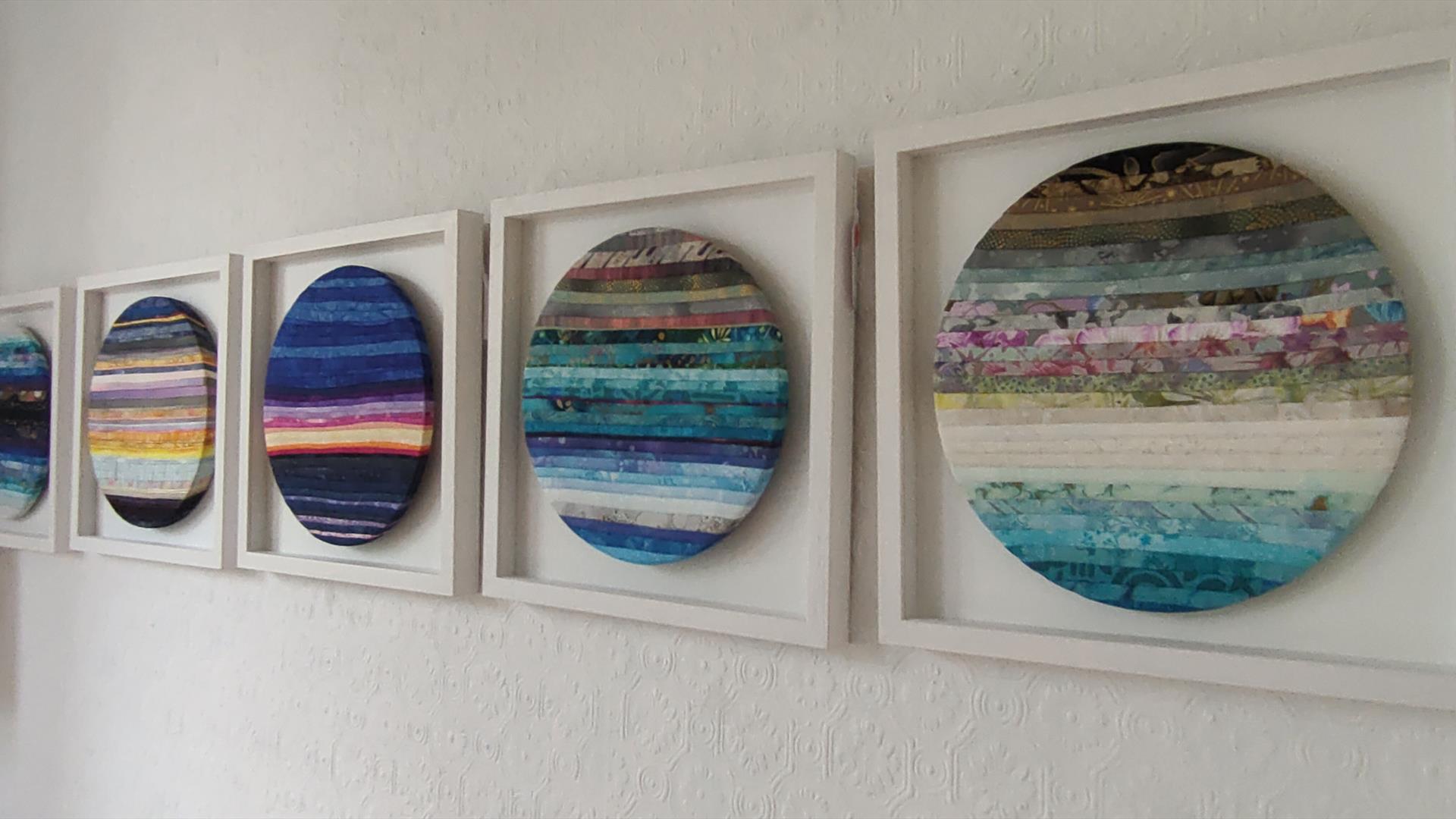 A row of five circular textile artworks in square white frames, showing layered horizontal bands of colour made from stitched fabric strips, arranged