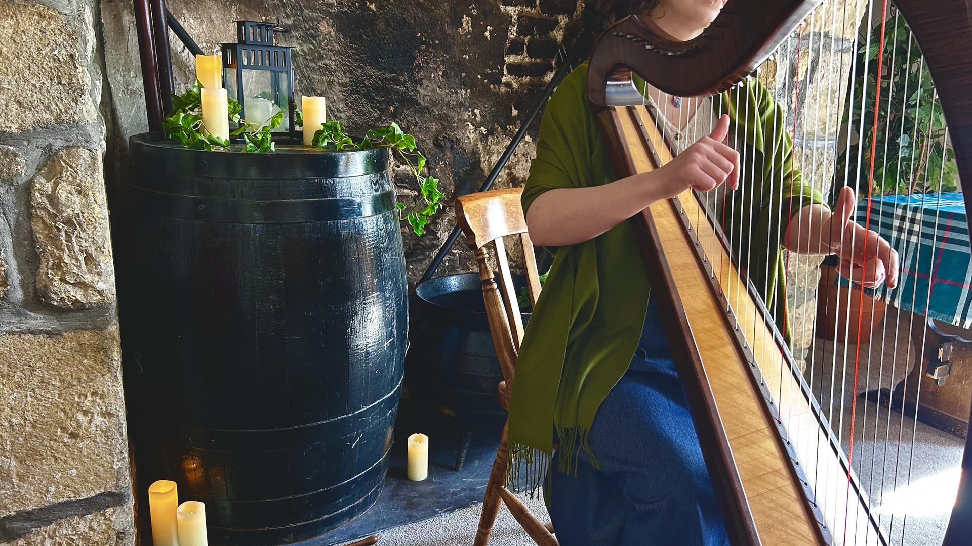 A musician plays a harp in a rustic room with stone walls, candlelight, and soft natural light