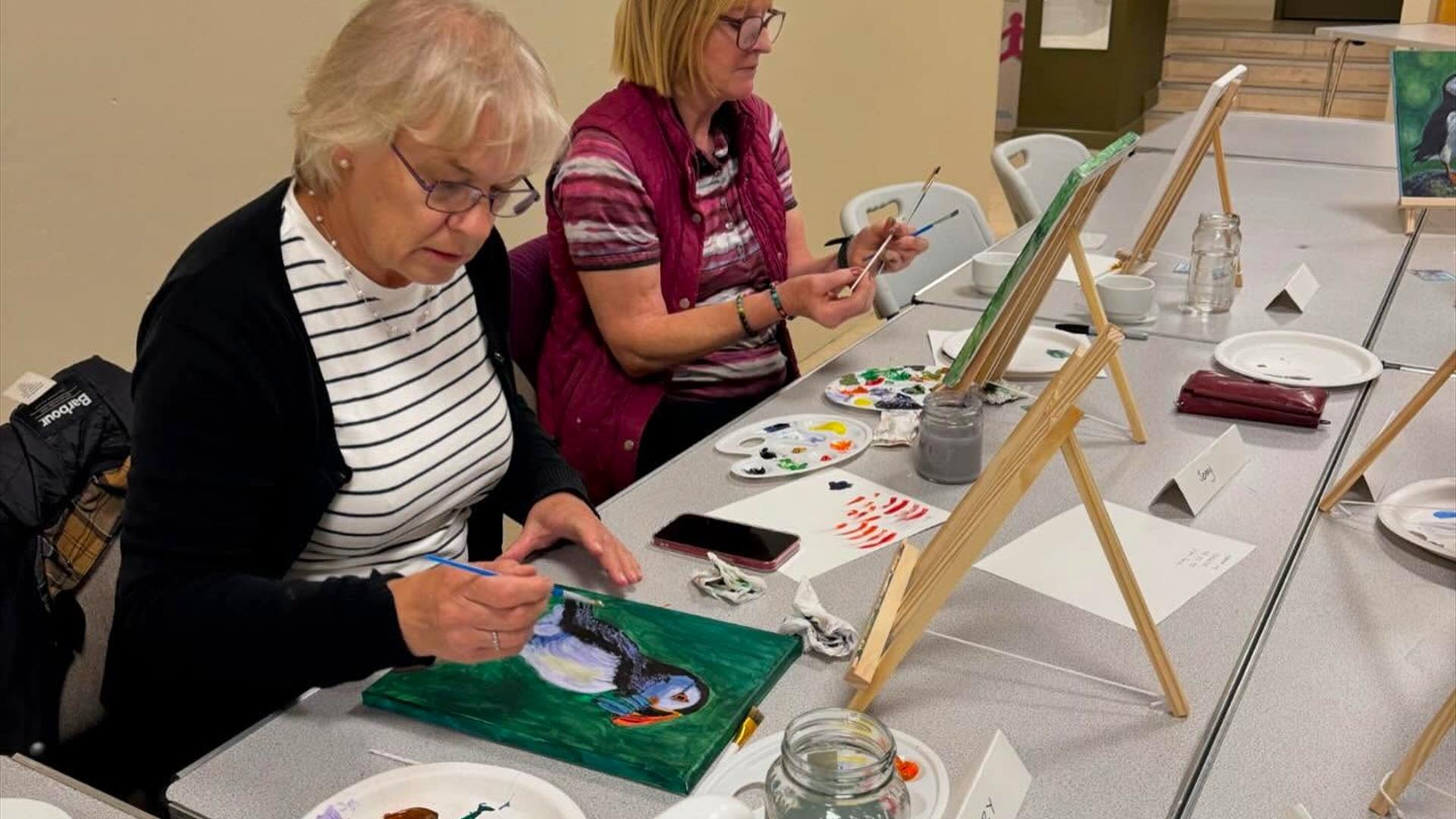 women sat in Midtown Makers painting on small canvas