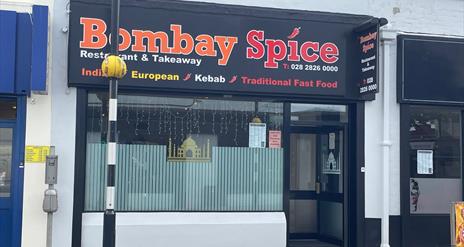 Exterior of Bombay Spice restaurant with pedestrian crossing in front of it and large window with entrance door.