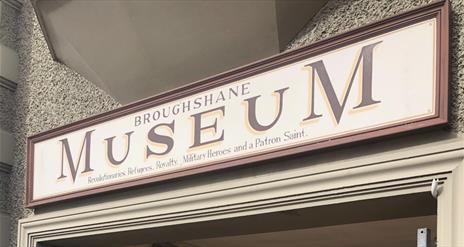 Red and Cream sign on building stating Broughshane Museum - Revolutionaries, Refugees, Royalty, Military Heroes and a Patron Saint.