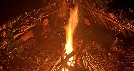 Two women laying under stick huts around a camp fire.