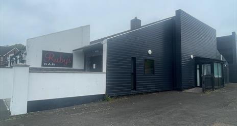 Black and white exterior of Ruby's Bar in Larne with sign featuring red name